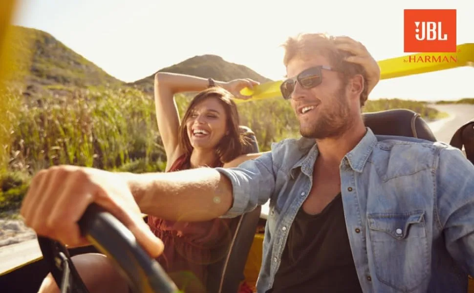 De tekst toont het 'JBL'-logo. Marketing lifestyle-foto van twee mensen in een auto met een zonovergoten buitenomgeving.